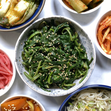 korean spinach in white bowl surrounded by assorted korean vegetable banchans: kimchi, oi kimchi, spicy marinated radish, bean sprouts, carrots, pickled daikon